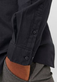 Camisa de un azul oscuro con puños abotonados y botones negros contrastantes. Textura suave con un ajuste clásico. Pantalones grises con patrón son parcialmente visibles.