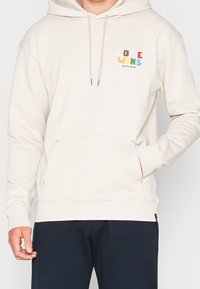 Felpa beige con tasca a marsupio. Presenta il testo colorato "LOVE WINS" e dettagli viola sui lacci e sulle cuciture laterali. Tessuto morbido.
