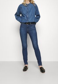 Camisa de mezclilla con botones y mangas abullonadas, combinada con jeans oscuros de corte slim y zapatos negros de charol tipo slip-on con un acento de hebilla dorada.