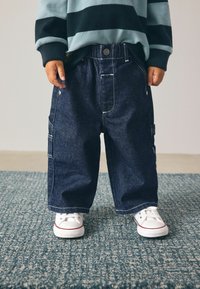 Pantalones cargo de mezclilla en azul oscuro con costuras blancas, que cuentan con bolsillos laterales y un corte relajado, combinados con zapatillas blancas.