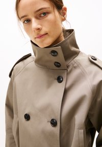 Trench-coat beige avec un col haut, design double boutonnage et boutons noirs. Tissu lisse avec épaulettes et poches latérales.
