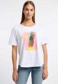 Witte katoenen T-shirt met een kleurrijke ananasafbeelding op een roze achtergrond met de tekst "FOLLOW THE SUN," gecombineerd met lichtblauwe jeans.