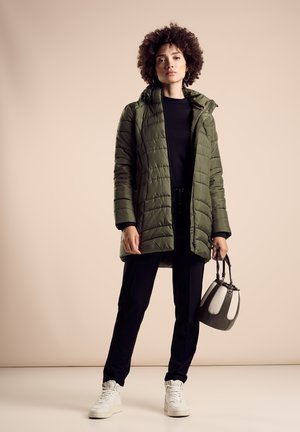 Manteau rembourré vert olive avec capuche, haut noir et pantalon noir. Baskets blanches et petit sac à main gris avec des accents clairs.