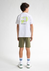 T-shirt en coton blanc avec un graphique vert vif au dos, associé à un short vert olive et des chaussures de sport blanches avec des accents verts.
