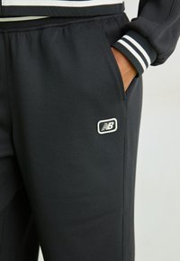 Pantaloni sportivi neri con finitura testurizzata, dotati di una tasca laterale e un logo New Balance delineato in bianco sulla coscia. Accenti a righe orizzontali.