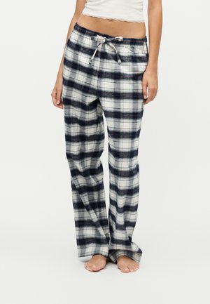 Femme portant un pantalon de pyjama ample à carreaux noirs et blancs avec une taille à cordon, debout pieds nus sur un fond uni.