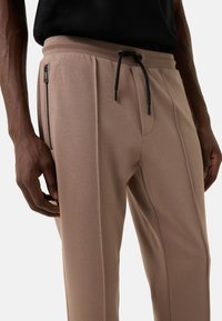 Beige joggerbroek met een tailleband met trekkoord, een zijzak met rits en subtiele plooien, gemaakt van een glad, zacht stof.