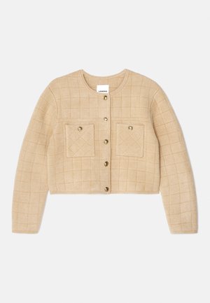 Μπεζ πλισέ καζάκα με στρογγυλή λαιμόκοψη, χρυσό κούμπωμα και δύο τσέπες στο στήθος. Μαλακή υφή με κομμένο μήκος.