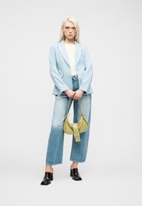 Γυναίκα με ξανθά μαλλιά που φοράει ανοιχτό μπλε σακάκι και τζιν, κρατώντας μια μικρή πράσινη τσάντα, στέκεται μπροστά σε απλό λευκό φόντο.