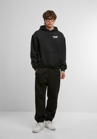 Black hoodie with a front pocket and white text logo, paired with matching black sweatpants and white sneakers. Smooth fabric texture.