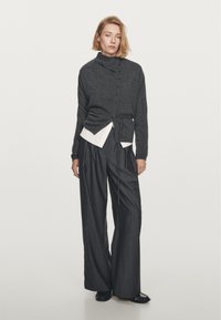 Kolaregrå button-up cardigan med hög krage, bärs över en vit skjorta, i kombination med löst sittande mörka skräddarsydda byxor. Slät textur.