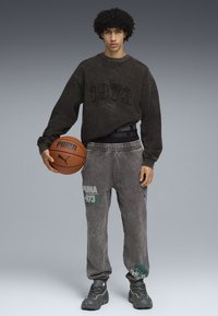 Felpa nera con il numero '1973' in rilievo e pantaloni sportivi grigi con testo verde e dettagli floreali. Sneakers grigie. Tiene un pallone da basket.