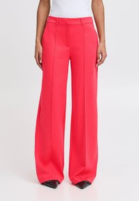 Pantalons larges de couleur corail vif, fabriqués en tissu lisse, présentant une taille haute, des plis nets et des poignets fuselés.