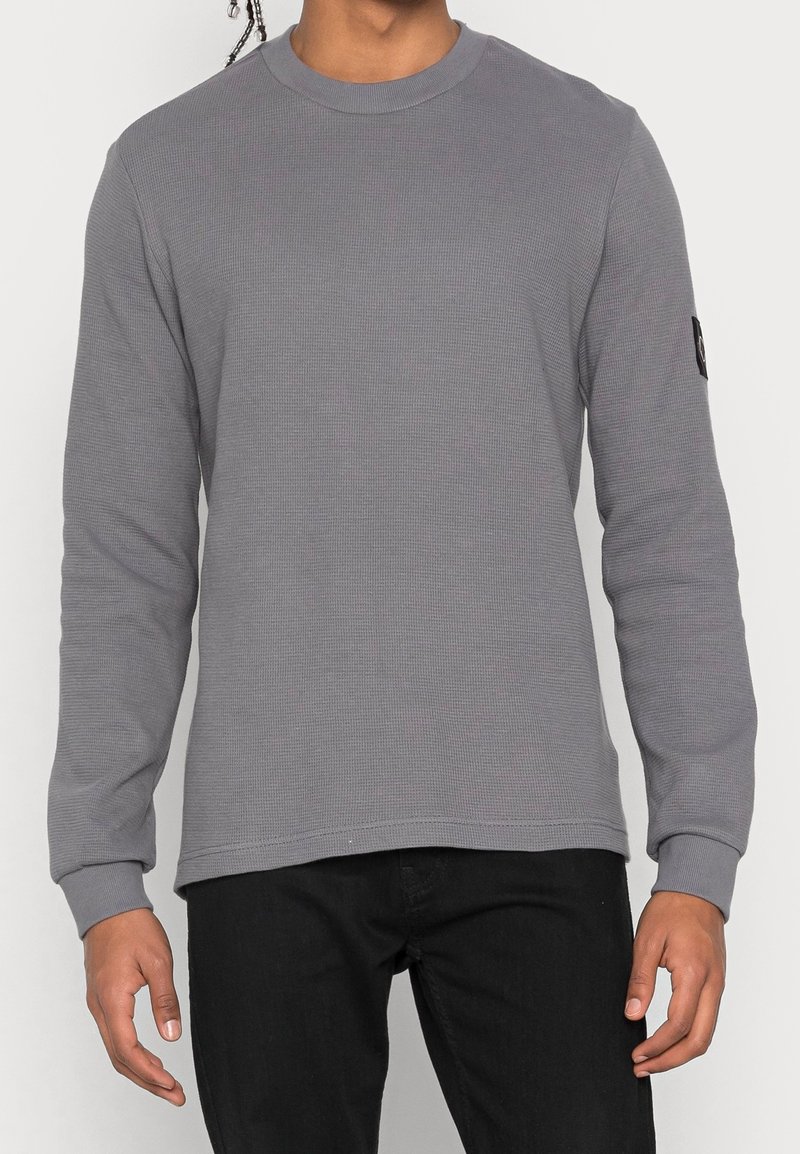 Grå långärmad tröja med texturerad tyg, rundad hals och ribbade manschetter. Har ett litet svart logomärke på vänster ärm.