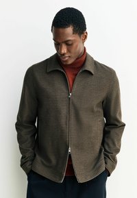 Braune Hahnentritt gemusterte Zip-up-Jacke aus einem strukturierten Stoff, mit Kragen und Seitentaschen, über einem kastanienbraunen Rollkragenpullover getragen.