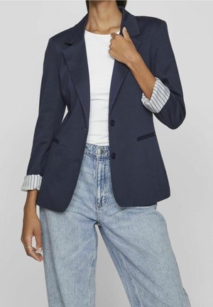 Kvinde iført en navy blazer med opkørte stribede manchetter, hvid skjorte og lyseblå højtsiddende jeans, poserer med den ene hånd nær brystet.