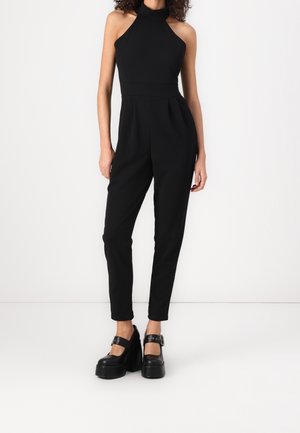 Sort ærmeløs jumpsuit med høj hals, tilspidsede ben og en tætsiddende talje. Kombineret med kraftige sorte plateau sko med spænder.