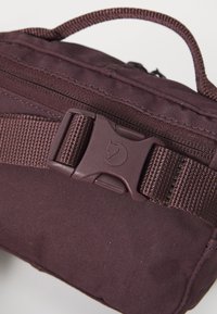 Tume burgundia kott, mis on valmistatud vastupidavast kangast, keskel on tõmblukk ja plastist pandla logo. Kaalub mugavaks kandmiseks pealmise käepideme.