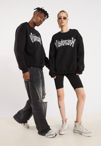 Sorte oversized hettegensere med "Visionary" i hvit skrift, kombinert med slitte grå jeans og svarte biker-shortser. Inkluderer hvite sneakers.