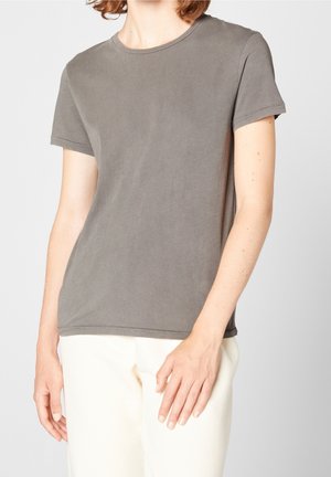 Osoba ubrana w gładki, szary t-shirt z krótkim rękawem i kremowe spodnie, stojąca na jednolitym jasnoszarym tle.