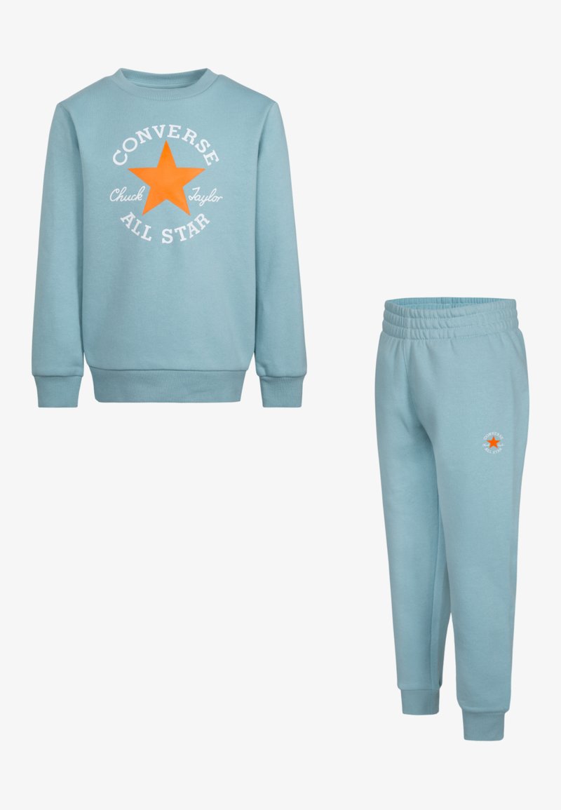 Ljusblå sweatshirt och matchande byxor i bomull, med en central orange stjärnlogotyp och vit text. Sweatshirten har ribbade ärmslut.