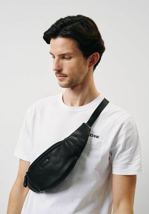 Czarna skórzana saszetka na biodra o zakrzywionym kształcie, z przednią kieszenią na zamek błyskawiczny oraz regulowanym paskiem noszonym na torsie na białej koszuli.