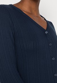 Nära bild på en mörkhyad person som bär en marinblå ribbad V-ringad topp med fyra knappar fram.