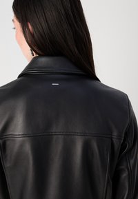 Een vrouw met lang, donker haar draagt een zwart leren jack, waarbij de details van de achterkant en de kraag van het jack goed zichtbaar zijn tegen een neutrale achtergrond.