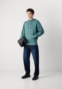 Felpa con cappuccio in cotone teal, caratterizzata da una tasca frontale e lacci; abbinata a jeans a gamba dritta blu scuro e scarpe nere.
