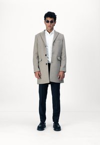 Beige Wollmantel mit Reverskragen, einem Knopfverschluss und seitlichen Taschen, getragen über einem weißen Hemd und schwarzen Hosen mit schwarzen Schuhen.