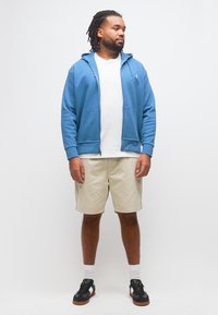 Άνδρας με κοτσίδες dreadlocks, φορώντας μπλε φούτερ με φερμουάρ, λευκό μπλουζάκι, μπεζ σορτς, λευκές κάλτσες και μαύρα αθλητικά παπούτσια, στέκεται μπροστά σε λευκό φόντο.