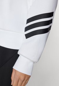 Hvit hettegensser med svart tre-stripes design på ermene. Mykt bomullsmateriale. Svart midjebånd og mansjetter. Enkel, minimalistisk estetikk.
