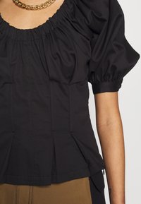 Schwarze Bluse mit Puffärmeln, gerafftem Ausschnitt, strukturiertem Oberteil, gefälteltem Saum und glatter Textur. Kombiniert mit braunen Hosen.