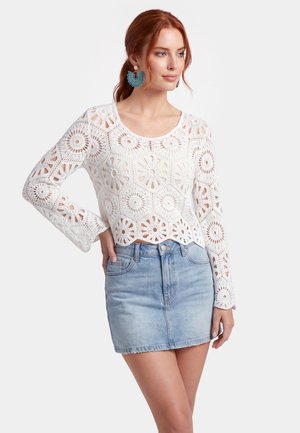 Donna con capelli rossi che indossa grandi orecchini blu, un top bianco lavorato a uncinetto a maniche lunghe e una gonna di jeans chiara, in piedi con una mano sul fianco.
