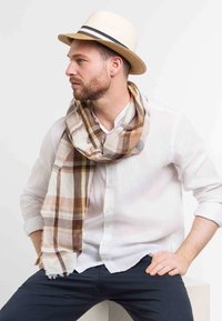 Bufanda a cuadros en tonos beige y marrón sobre una camisa blanca, combinada con un sombrero de paja claro con una cinta negra.