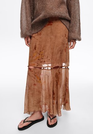 Brązowa, przezroczysta długa spódnica w kwiaty z koralikami na wysokości kolan, zestawiona z puszystym brązowym sweterkiem oraz czarnymi sandałami na palce ozdobionymi koralikami na modelce.