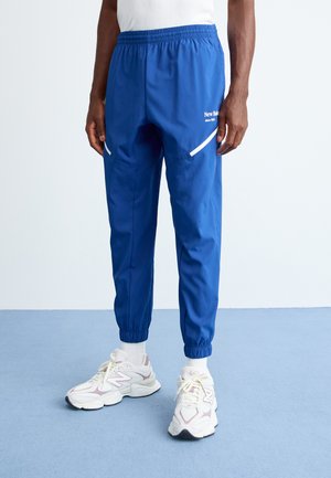Person wearing blue New Balance track pants with white reflective details and white sneakers standing on a blue floor.