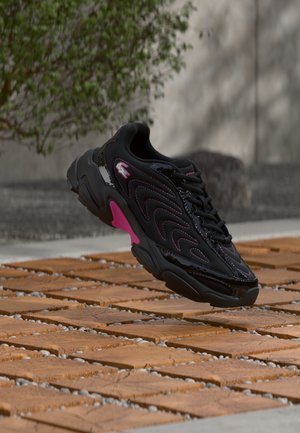 Črno-roza superga z vzorcem valov in logotipom, obešena nad leseno in prodnato zunanjo površino z zamegljenim zelenjem in ozadjem z zidom.