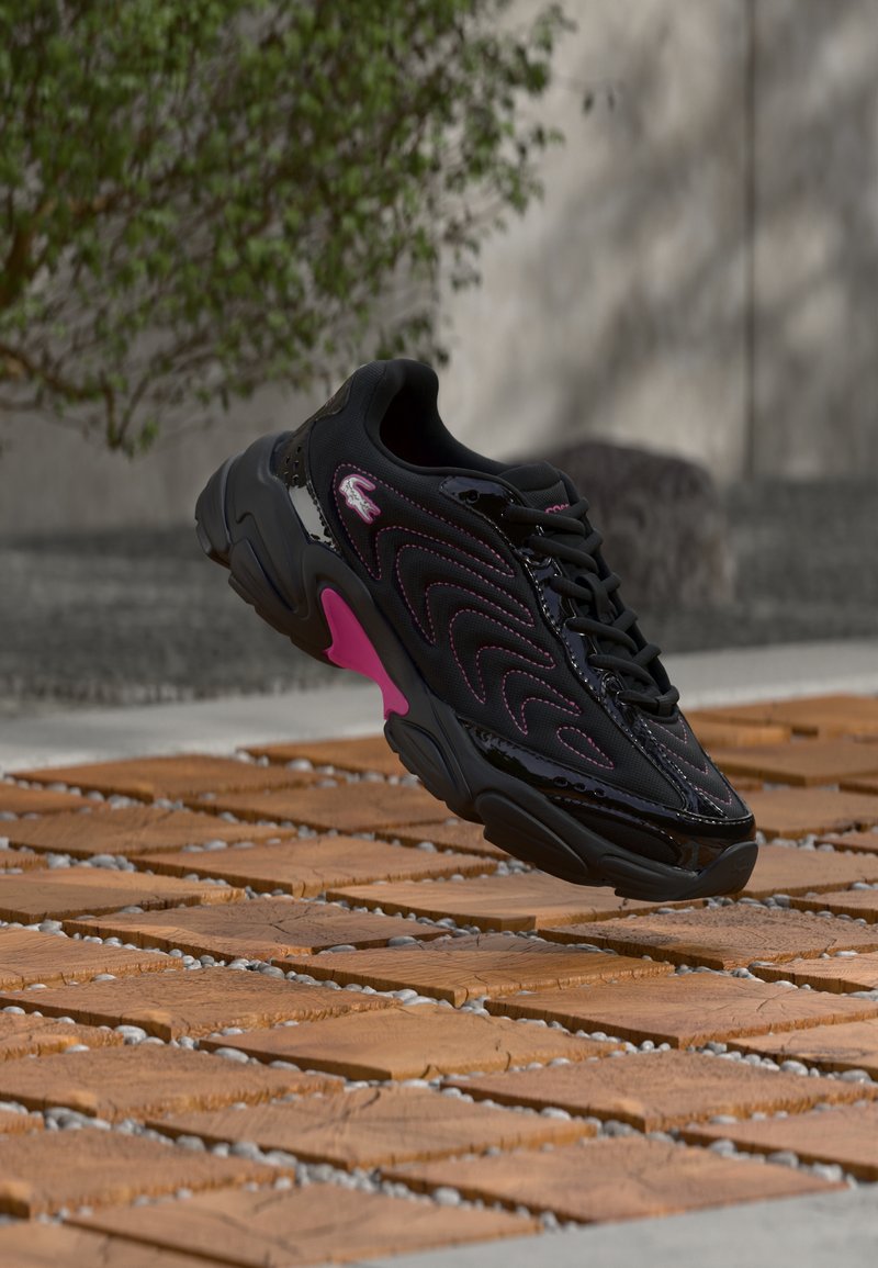 Μαύρο και ροζ αθλητικό παπούτσι με κυματιστό σχέδιο και λογότυπο, κρεμασμένο πάνω από ξύλινη επιφάνεια και χαλίκια σε εξωτερικό χώρο με θολό φόντο πράσινου και τοιχοποιίας.