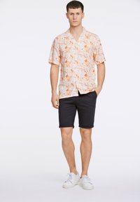 Camisa de flores de manga corta en color crema con patrones naranjas, combinada con pantalones cortos azul marino. El modelo lleva zapatillas blancas, mostrando un look casual.