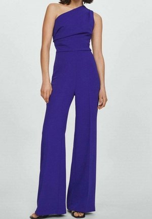 Femme portant une combinaison bleu roi sans manches à une épaule, pantalon à jambes larges et sandales noires à bouts ouverts, debout devant un fond uni.