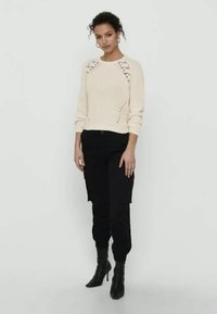 Cremig gerippter Strickpullover mit Schnürdetails an den Schultern, kombiniert mit schwarzen Cargo-Hosen und schwarzen High-Heels. Schlicher Hintergrund.