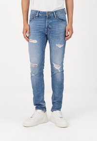 Világoskék, kopott farmernadrág, amelyen szakadt részek és rojtos szélek találhatók, slim fit kialakítással és öt zsebbel. Fehér sportcipővel párosítva.
