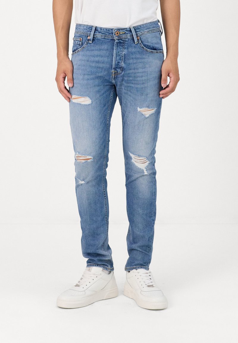 Világoskék, kopott farmernadrág, amelyen szakadt részek és rojtos szélek találhatók, slim fit kialakítással és öt zsebbel. Fehér sportcipővel párosítva.