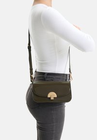 Kreuzkörpertasche aus olivgrünem Lackleder mit gebogenem Verschluss. Verfügt über einen langen verstellbaren Riemen und ein schlankes rechteckiges Design.