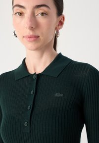 Temno zelena rebrasta polo srajca s klasičnim ovratnikom, ki ima pet gumbov in majhen, izšit logotip Lacoste na prsih.