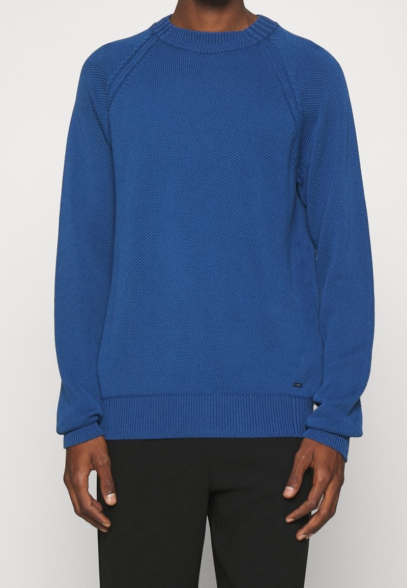 Personne portant un pull bleu texturé à manches longues et un pantalon noir, debout contre un fond clair uni.