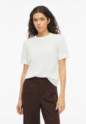 Femme aux longs cheveux bouclés portant un haut blanc texturé à manches courtes et un pantalon taille haute sombre, debout devant un fond blanc uni.