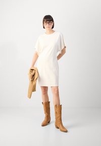 Krem haljina kratkih rukava s širokim rukavima, uparena s smeškim kožnim čizmama do kolena. Manekenka drži odgovarajuću smeđu jaknu. Minimalistički dizajn.