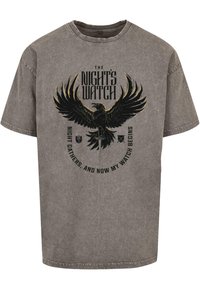Grijze katoenen T-shirt met een zwarte adelaar afdruk en de tekst "THE NIGHT'S WATCH" naast een zwaard, tegen een gestructureerde achtergrond.